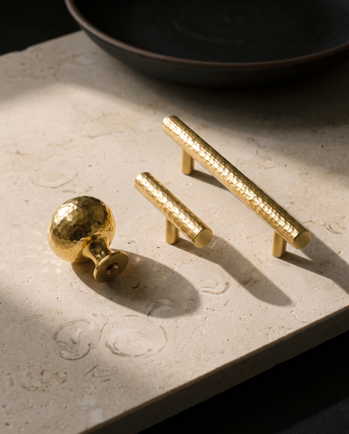 The Atelier hammered brass knob, T bar and pull laid out on honed cream limestone in raking afternoon window light