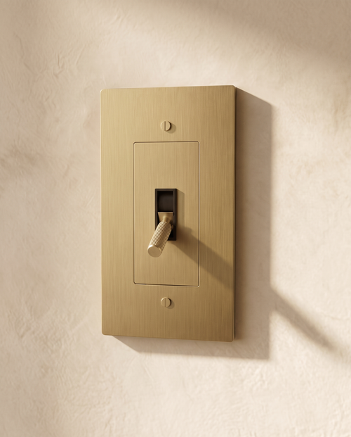 Brio Brass Toggle Switch in brushed brass, packshot on warm ivory plaster