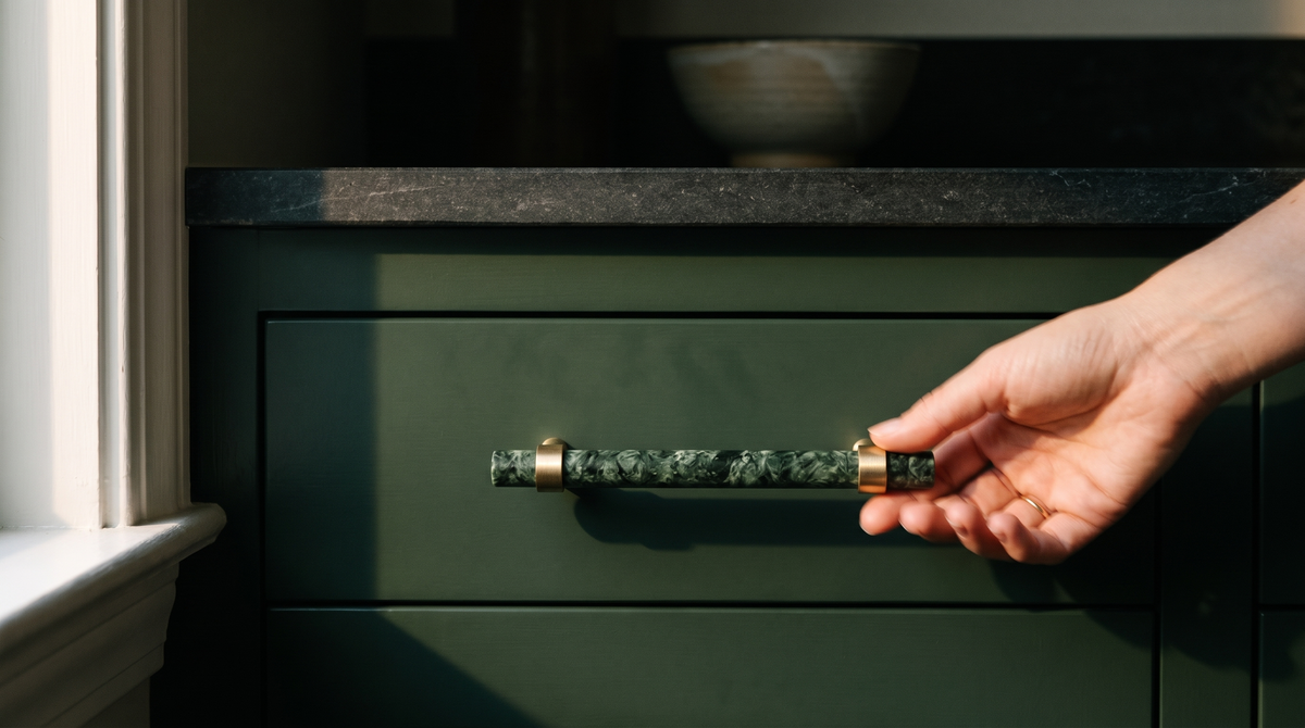 A hand reaching for a Verde green marble and brass pull on a deep green kitchen drawer in late afternoon light